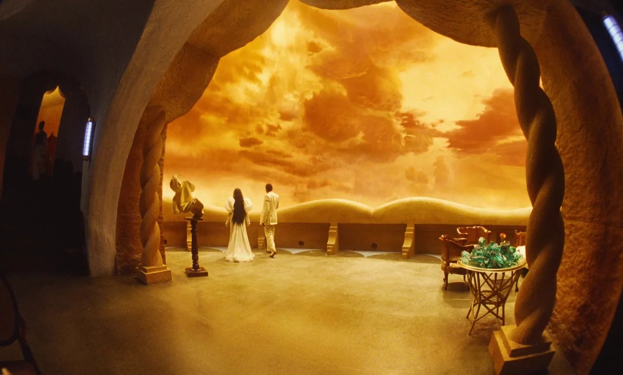 Poor Things (2023) movie still: wide — A wide interior space features a large, curved, luminous orange wall resembling a sunset. A man and a woman…