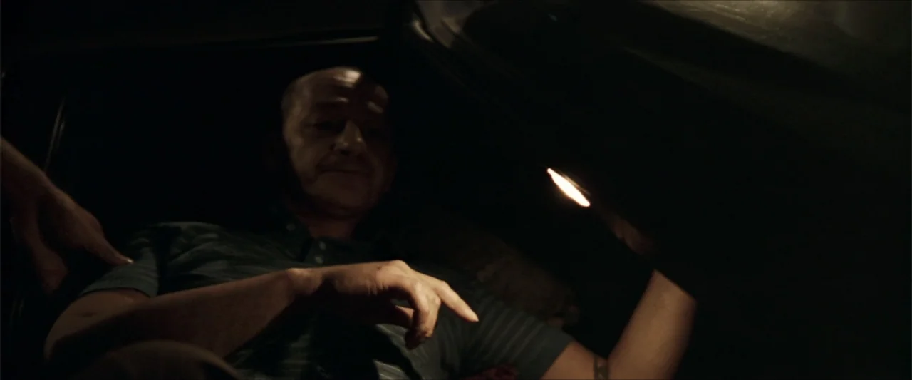 GOMORRAH (2023) movie still: medium — The interior of a vehicle is dark except for a small overhead light source. A bald middle-aged man sits in the…