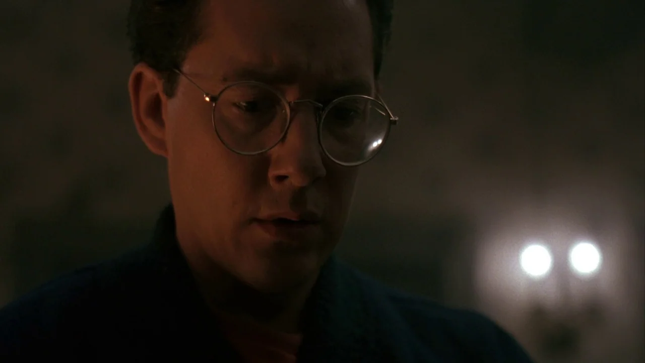 Jason Goes To Hell (1993) movie still: closeup — A dimly lit room contains a man in glasses looking downward. The background is mostly obscured by…