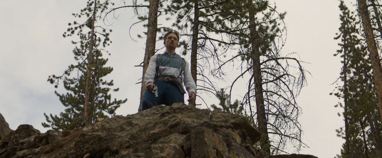 Damsel (2018) movie still: fullbody — A man stands atop a rugged rock formation surrounded by tall, thin coniferous trees. He looks downward while…
