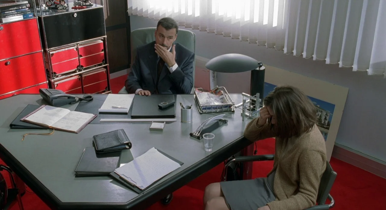 Read My Lips (2001) movie still: wide — A modern office features a large angular grey desk, red carpeting, and red shelving units. A man sits behind…