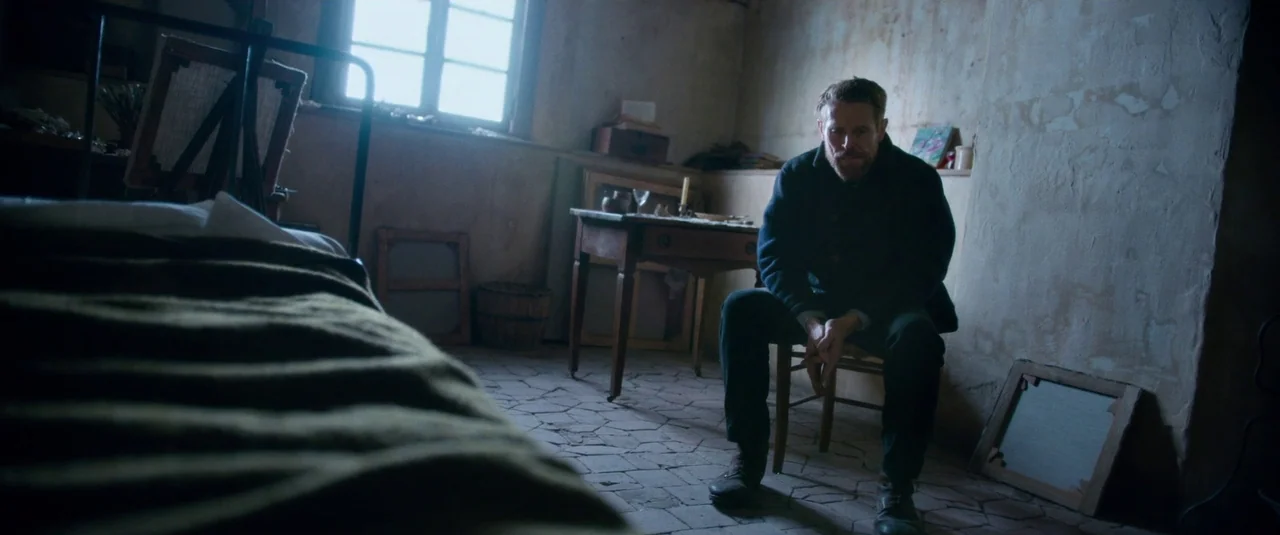 At Eternity’s Gate (2018) movie still: medium — A sparsely furnished, dimly lit room with rough plastered walls and a tiled floor. A man sits…