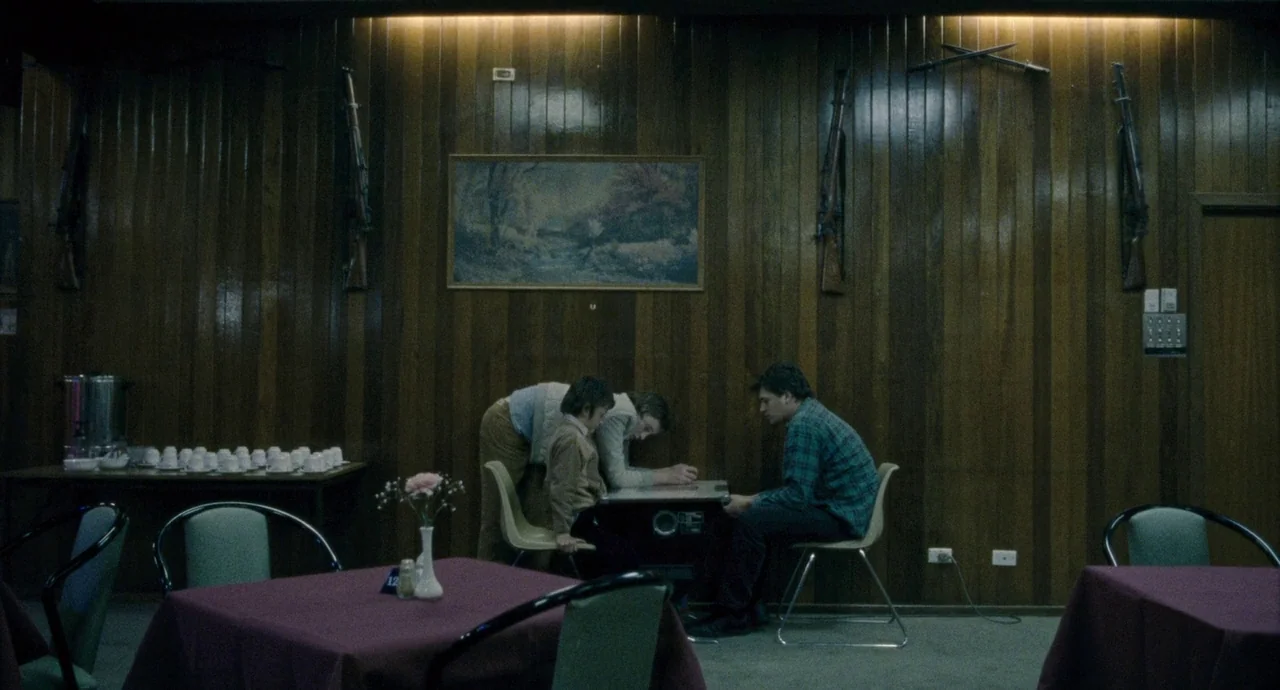 Snowtown (2011) movie still: wide — A community hall features dark vertical wood paneling decorated with antique rifles and paintings. Three figures…