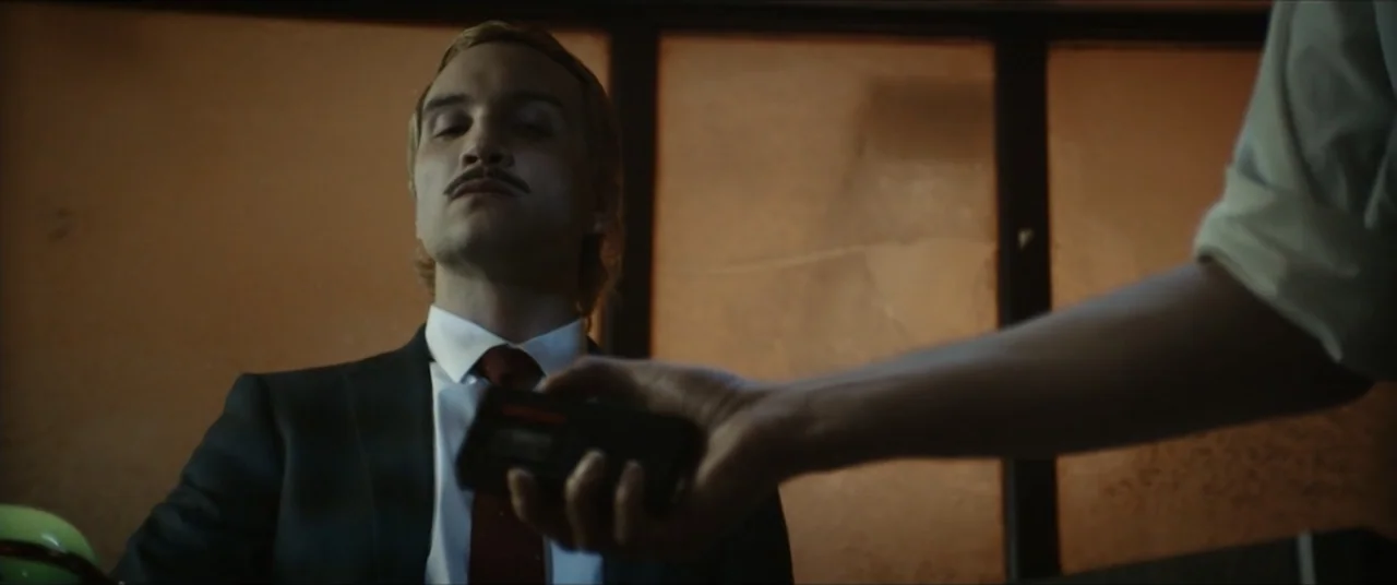 Vicious Fun (2021) movie still: medium — A dark, wood-paneled room serves as the setting for an exchange. A man in a suit looks ahead while a…