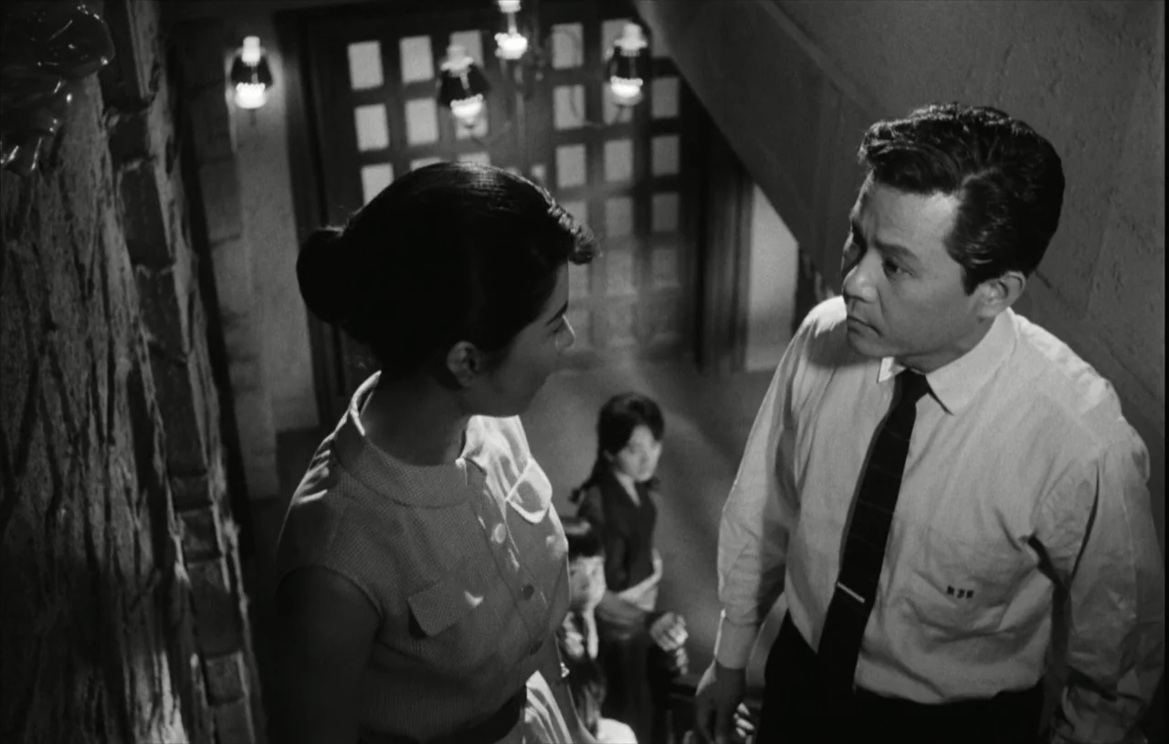 The Housemaid (1960) movie still: medium — A stone staircase in an interior space with sconce lights mounted on the wall. A man and a woman face one…