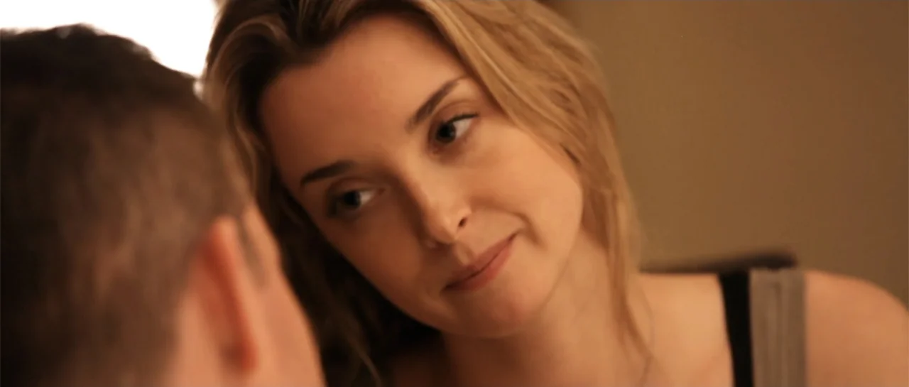 La coherencia del caos movie still: closeup — An interior space provides a neutral background for the two subjects. A woman in a grey tank top looks…