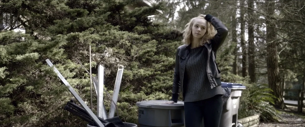 The Hollow Child (2018) movie still: medium — A residential yard with dense evergreen shrubs and several gray plastic trash cans. A young woman…