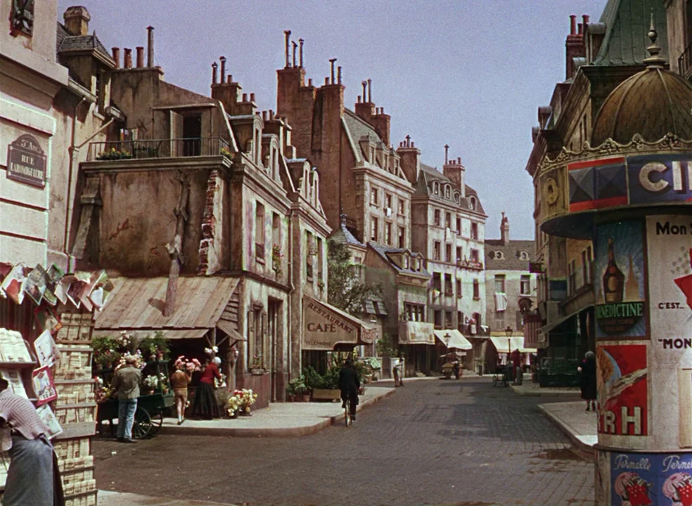 An American In Paris (1951) movie still: establishing — A Paris street with multistory stone buildings, tall chimneys, and a Morris column kiosk.…