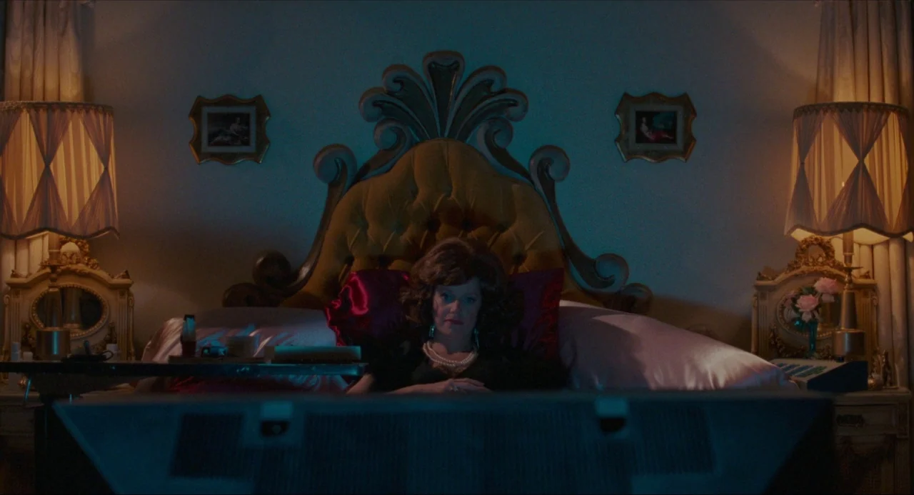 True Stories (1986) movie still: medium — A dimly lit hotel room with a prominent yellow velvet headboard and dual lamps. A woman rests against the…