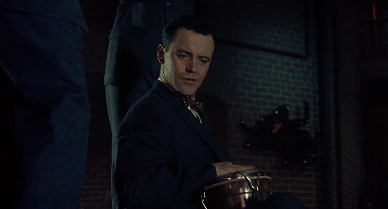 Bell Book and Candle (1958) movie still: medium — An outdoor brick wall at night serves as the backdrop for the musician. A man sits in a dark suit…