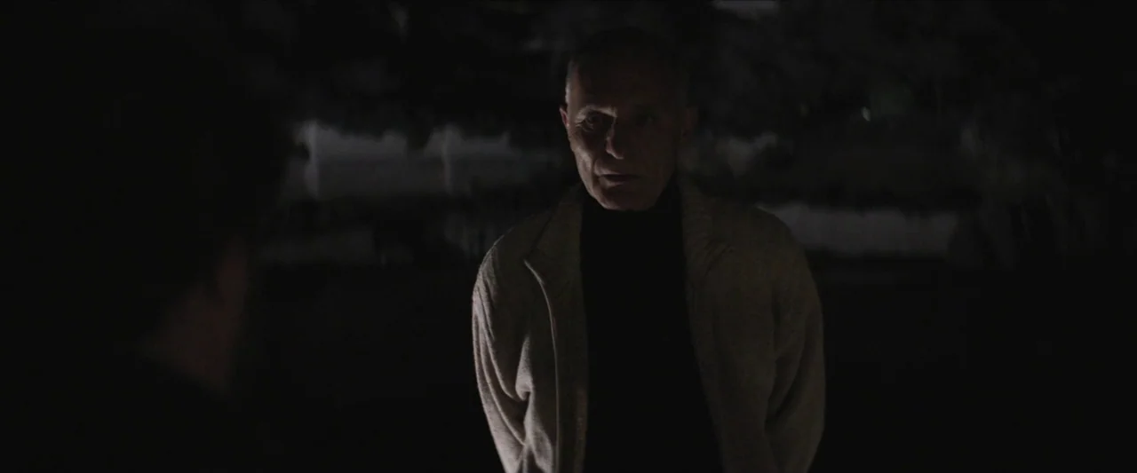 Chevalier (2015) movie still: medium — An older man stands outdoors at night in a dark, indistinct park environment. He wears a beige knit cardigan…