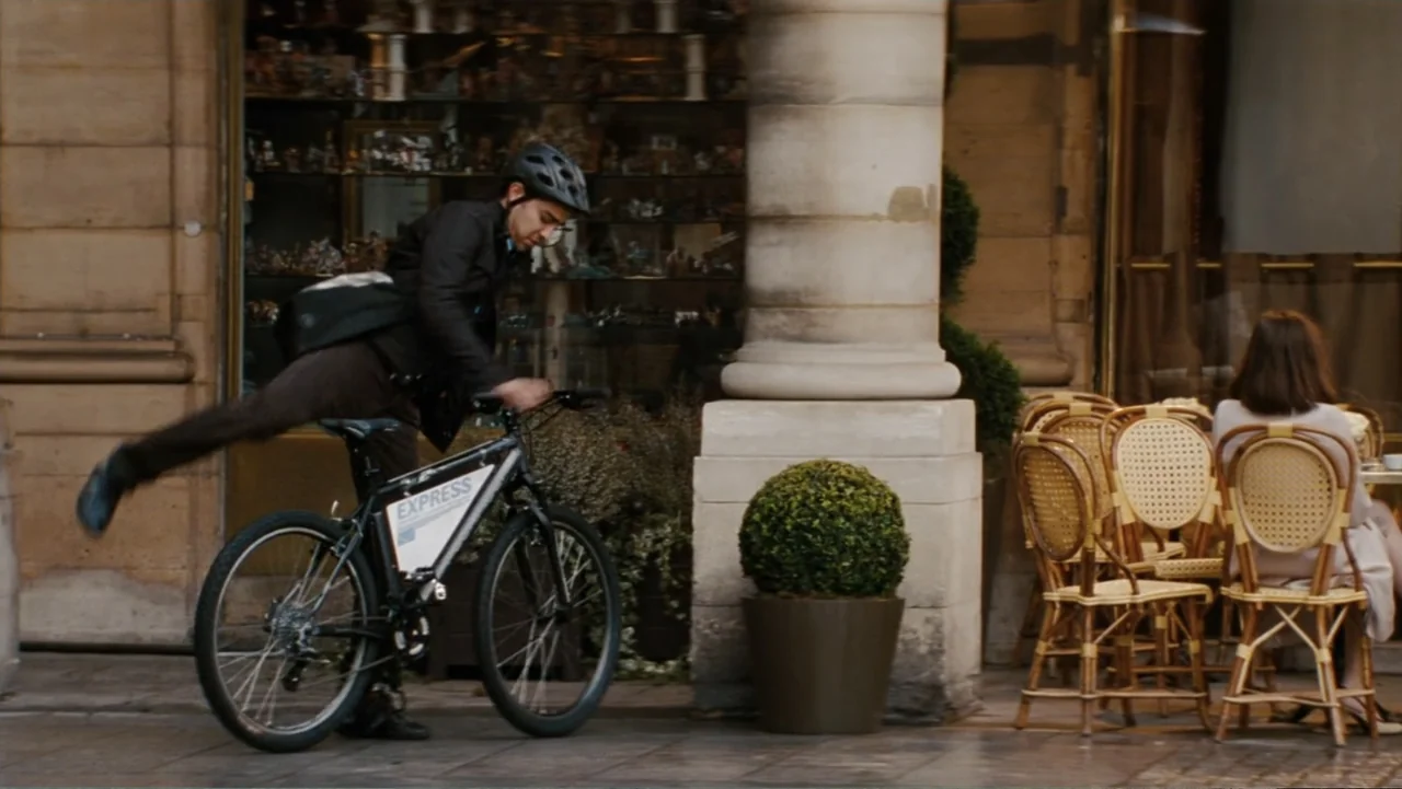 The Tourist (2010) movie still: fullbody — A Parisian street corner features stone pillars and a sidewalk cafe with wicker chairs. A courier rides a…