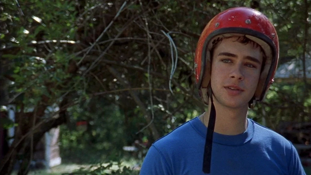 Mean Creek (2004) movie still: medium — A teenage male stands in a wooded area with dense greenery behind him. He wears a bright red motorcycle…