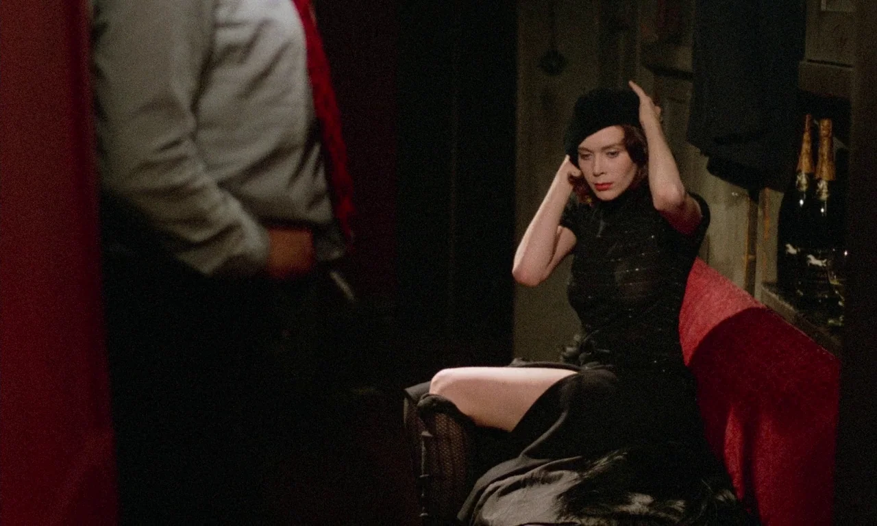 The Margin (1976) movie still: medium — A dark booth features deep red velvet upholstery and bottles placed in the shadows. A woman sits on the edge…