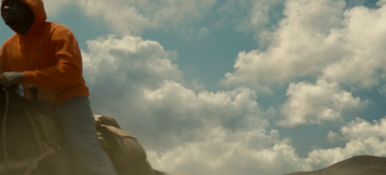 Nope (2022) movie still: medium — A person sits on horseback under a vast, cloud-filled bright blue sky. The frame focuses on the upper body of the…