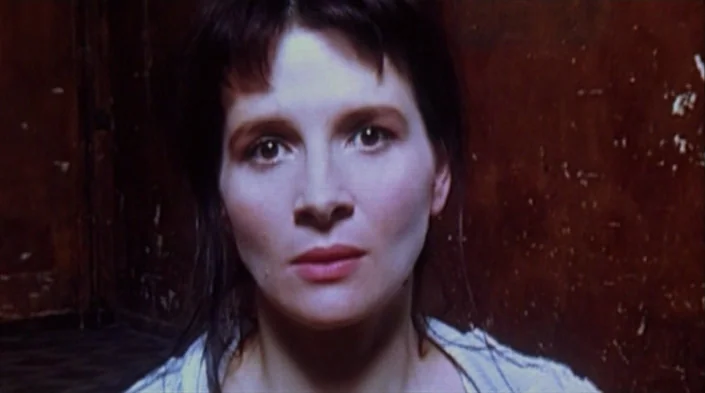 Code Unknown (2000) movie still: closeup — A woman stands before a dark wall with weathered, peeling dark red paint. She faces forward while looking…