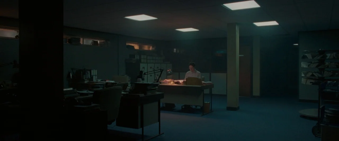 Censor (2021) movie still: wide — A dimly lit, expansive open-plan office features rows of desks and metal filing cabinets. A lone individual works…