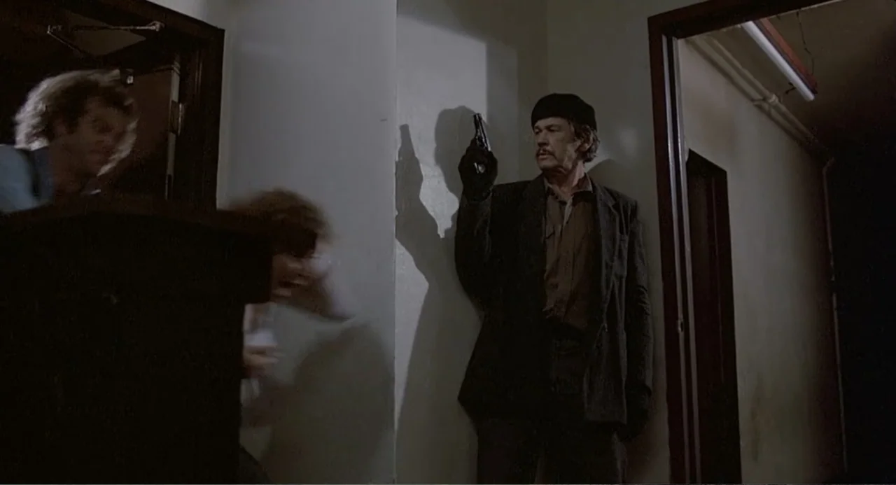 Death Wish II (1982) movie still: medium — A dimly lit indoor hallway features plain walls and dark wooden door frames. An older man points a…