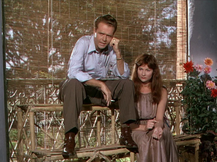 The River (1951) movie still: medium — A man and a woman sit on a bamboo bench behind a woven reed partition. Flowers are visible in the foreground…