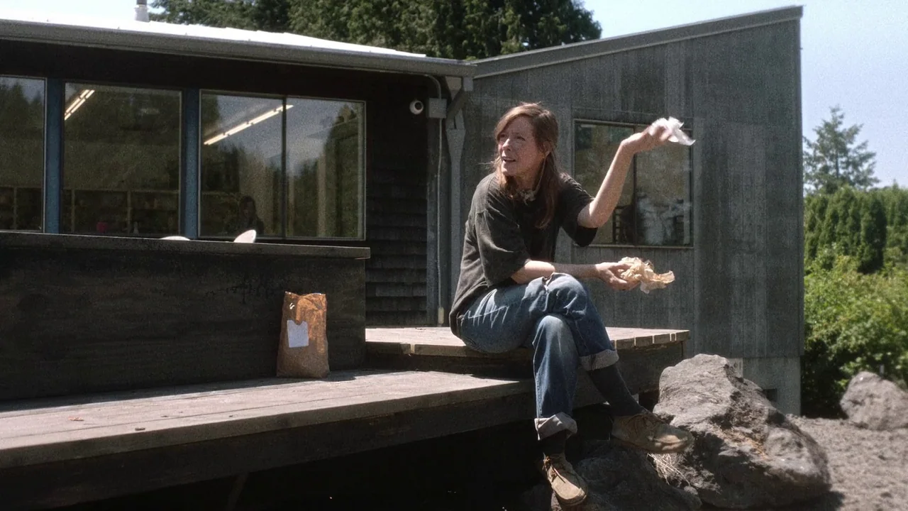 Showing Up (2022) movie still: medium — A woman sits on a wooden deck outside a modern building with gray siding. She holds a small paper bag while…