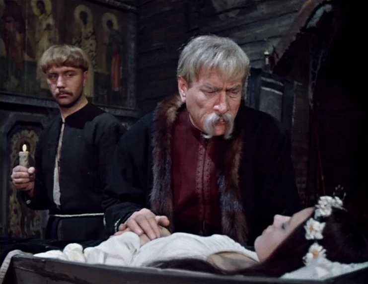 Viy (1967) movie still: medium — A dark wooden chapel features religious icons mounted on the walls behind the subjects. An older man stands over a…