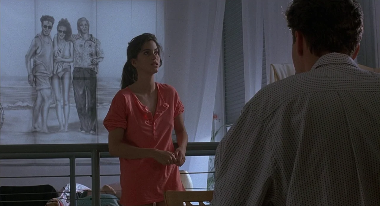 Less Than Zero (1987) movie still: medium — A sunlit apartment features a large wall mural depicting three people on a beach. A woman stands by a…