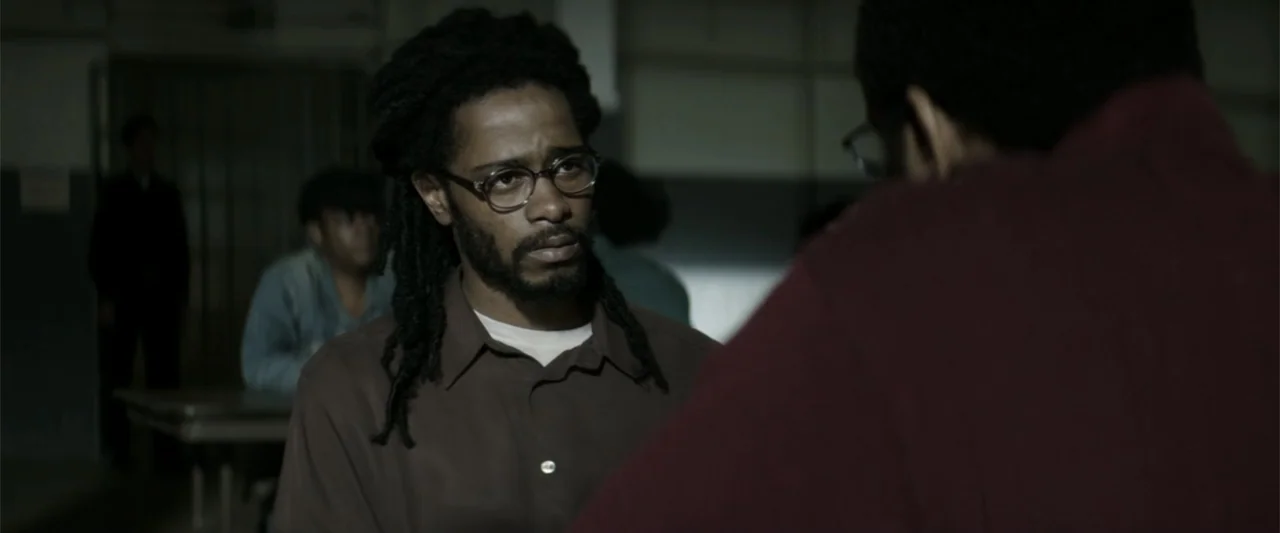 Crown Heights (2017) movie still: medium — A utilitarian room with blurred figures in the background and neutral wall tones. A man with dreadlocks…
