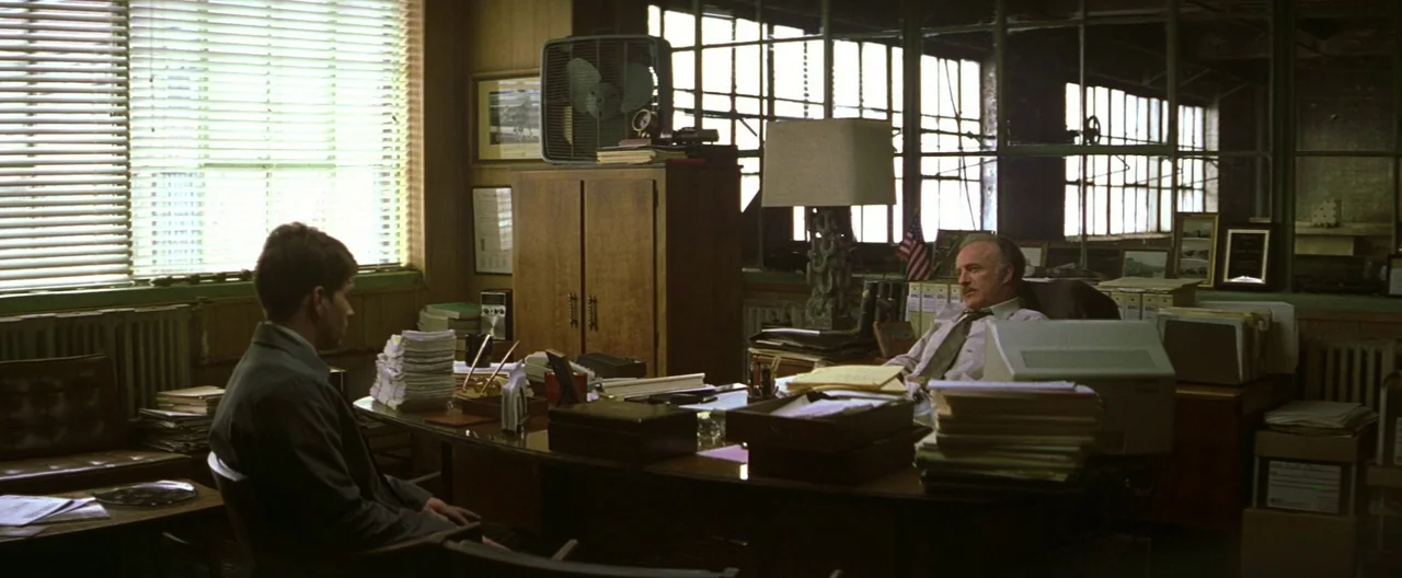 The Yards (2000) movie still: wide — A cluttered 1990s corporate office features wood grain furniture, stacks of files, and windows. Two men conduct…