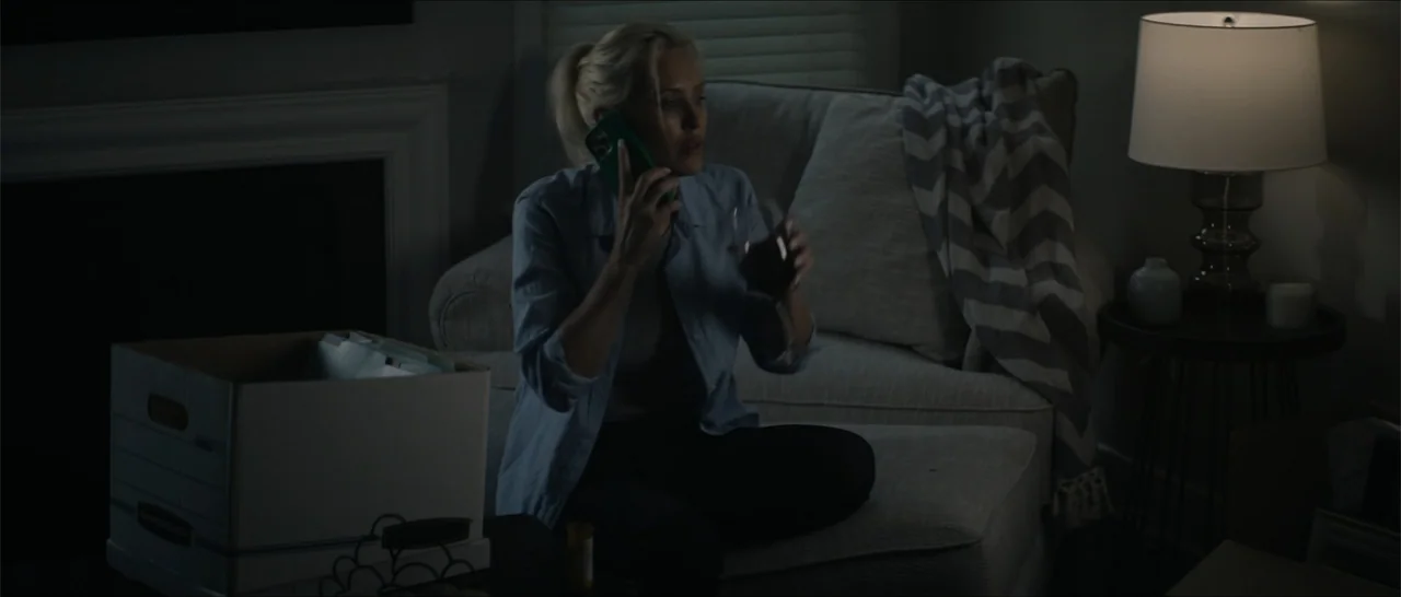 Crescent City (2024) movie still: medium — A dimly lit living room contains a sofa and a side table with a lamp. A woman sits on the sofa while…