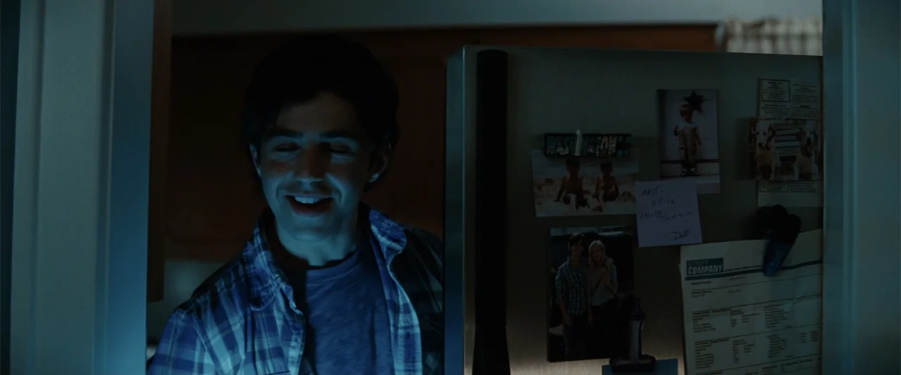 Red Dawn (2012) movie still: medium — A dark residential interior featuring a stainless steel refrigerator covered with various magnets and notes. A…