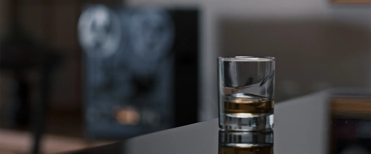 To Hear the Birds Singing (1995) movie still: closeup — A glass of amber liquid rests on a dark surface in a dimly lit room. A blurred reel-to-reel…