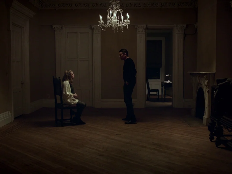 First Reformed (2017) movie still: wide — A spacious room with traditional crown molding and dark wooden floors. A woman sits on a wooden chair…