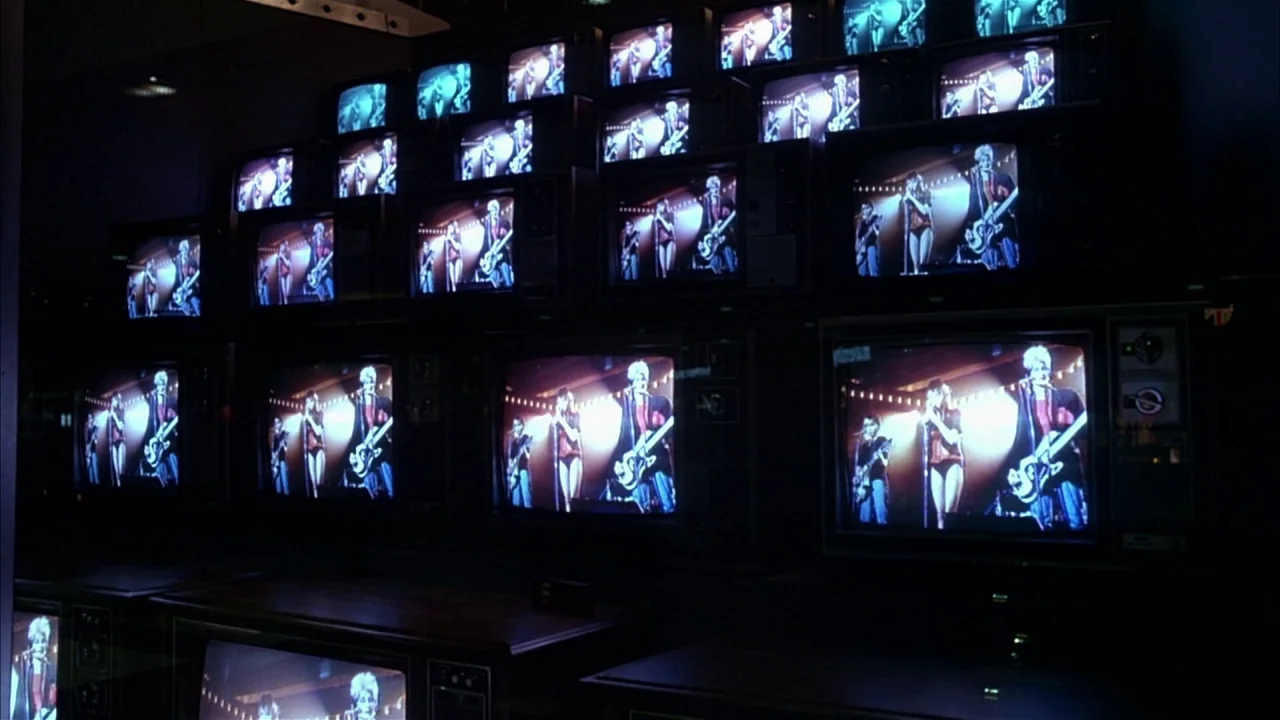 Ladies and Gentlemen, the Fabulous Stains (1985) movie still: wide — A dark retail space displays a large wall of stacked vintage television sets.…
