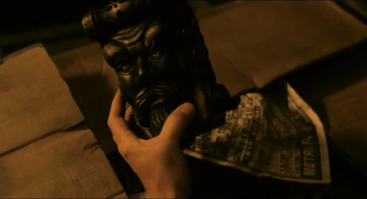 From Dusk Till Dawn (1996) movie still: closeup — A dark indoor space contains a piece of paper and a sculptural object. A hand enters the frame to…