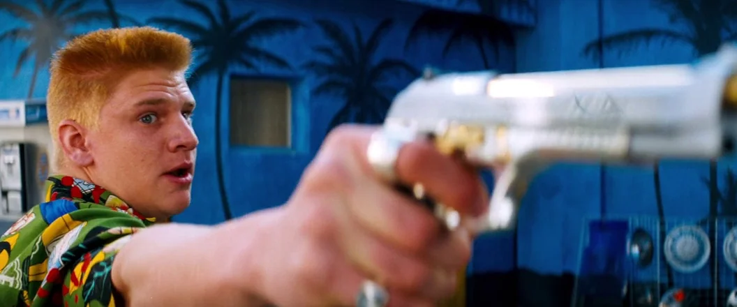 Romeo + Juliet (1996) movie still: medium — A man with a flat-top haircut stands in front of a bright blue wall. He points a metallic handgun toward…