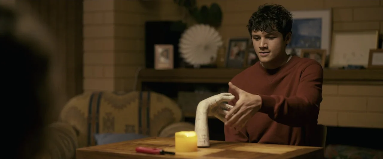 Talk to Me (2023) movie still: medium — A dimly lit room features a wooden table with a candle and an anatomical sculpture. A young man sits in the…