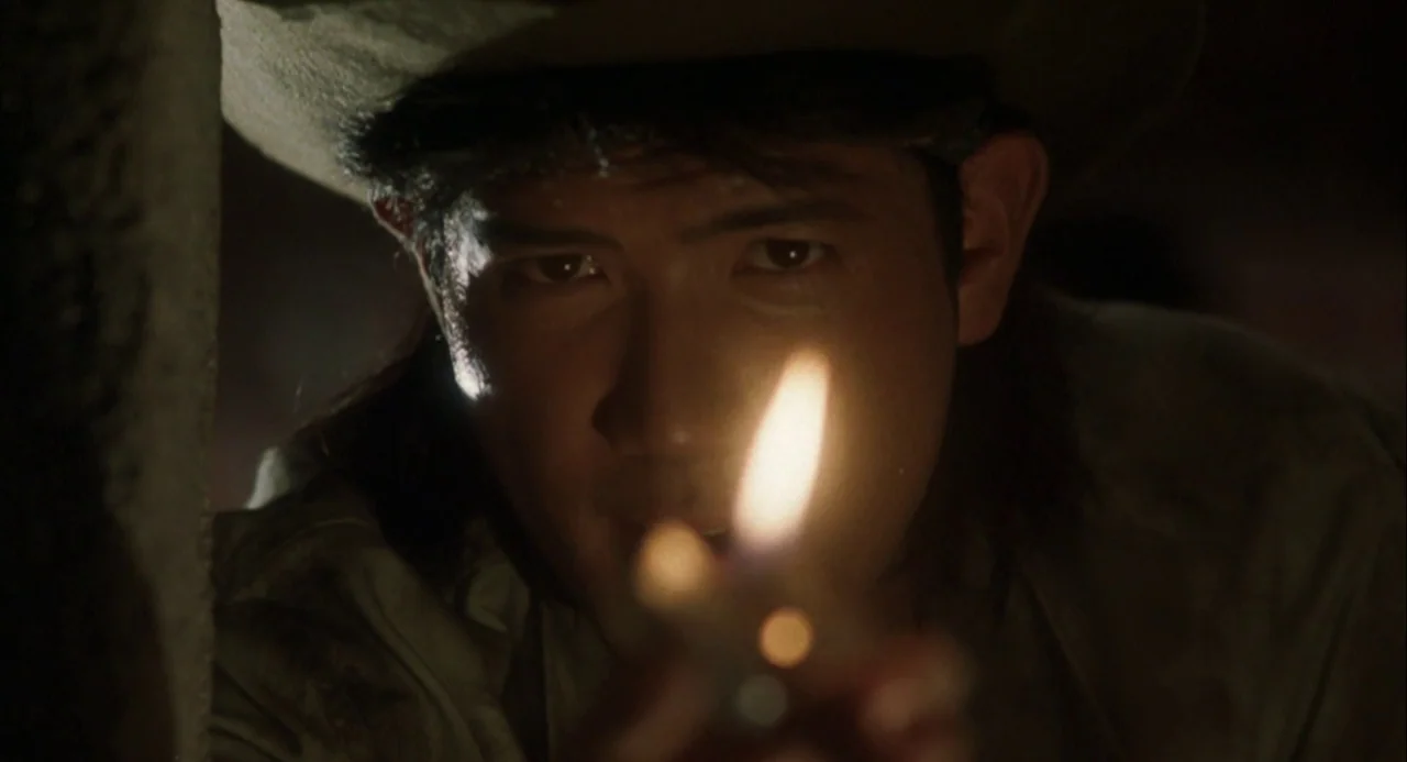 Godzilla and Mothra: The Battle for Earth (1992) movie still: closeup — A man in a brimmed hat sits in a dark, poorly lit environment. He holds a…