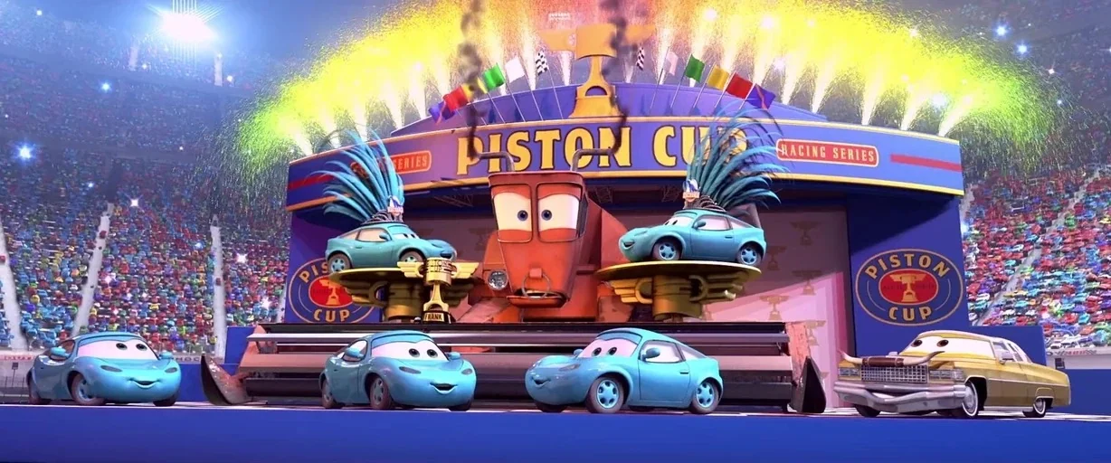 Cars (2006) movie still: wide — A grand stage at a racing stadium features large Piston Cup branding and pyrotechnics. Several anthropomorphic…