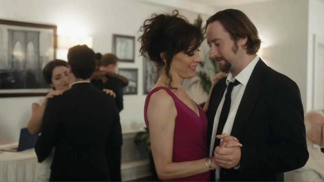 Toss It (2019) movie still: medium — A well-lit indoor venue features white walls decorated with framed black and white photos. A man in a suit and…