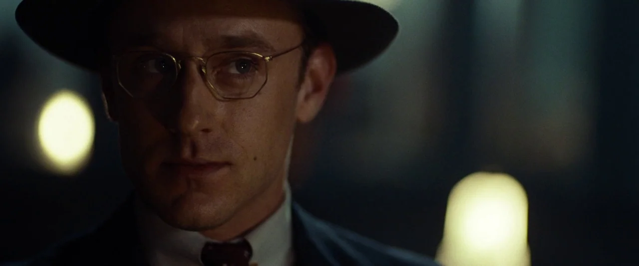 Kill Your Darlings (2013) movie still: closeup — A man stands in a dark room with out-of-focus light sources behind him. He wears a fedora and…