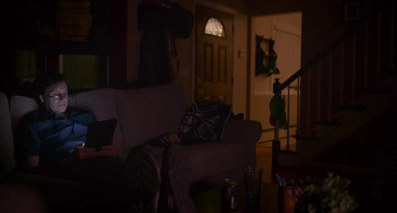 Eighth Grade (2018) movie still: medium — A dark, dimly lit domestic space features a person sitting on a cushioned sofa. The man is reading from a…