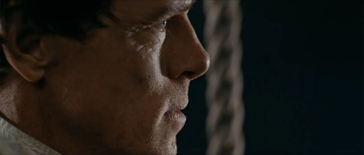 Out of Liberty (2019) movie still: closeup — A dark interior setting with a vertical rope visible in the blurred background. A middle-aged man in a…