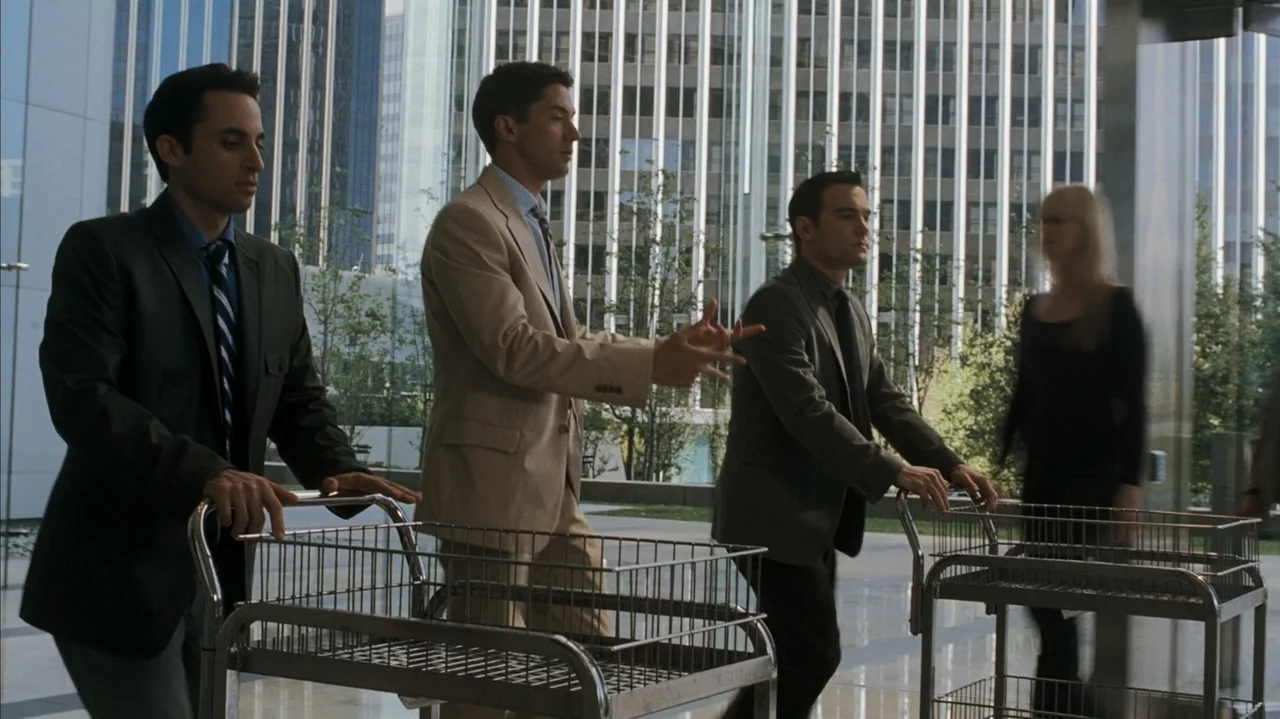 F Valentine's Day (2026) movie still: medium — Modern office building glass lobby with tall windows reflecting a skyscraper exterior. Three men in…