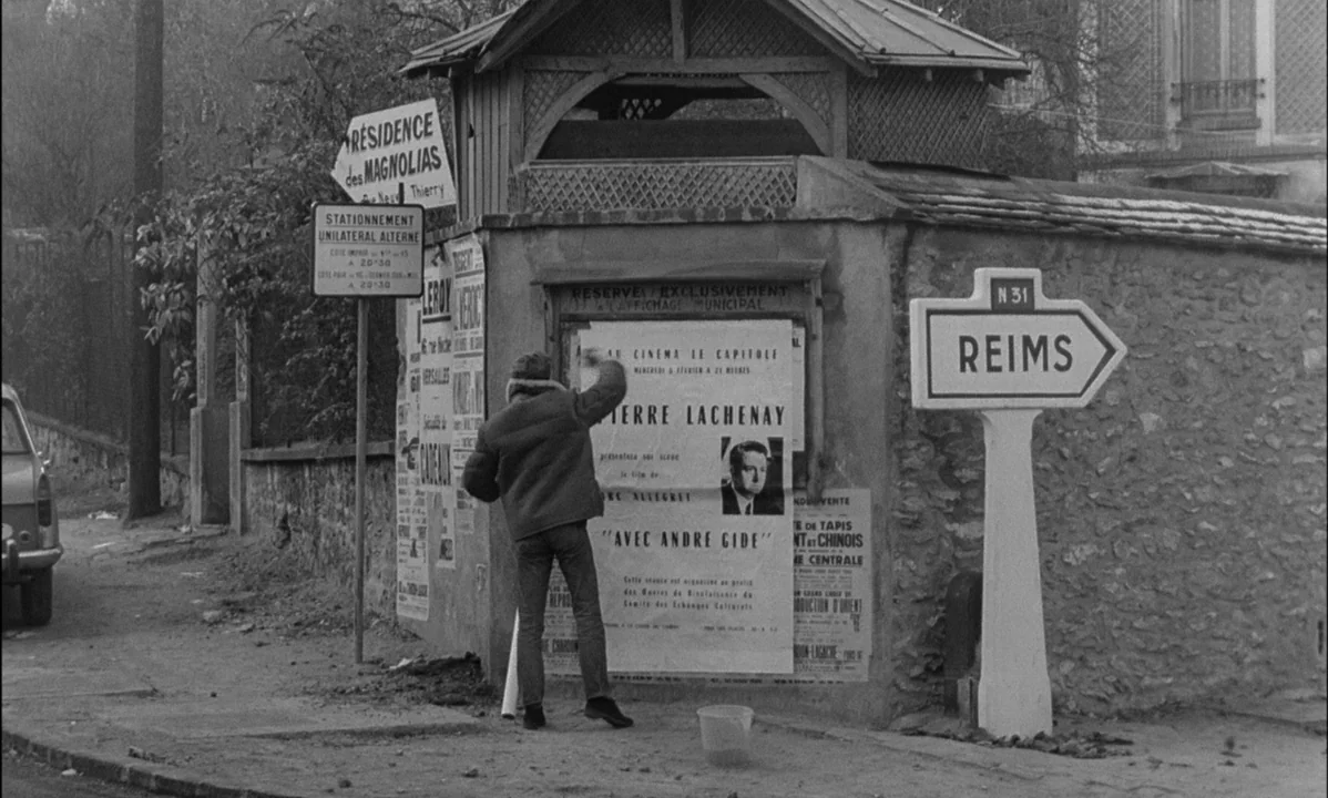La Peau Douce (The Soft Skin) (1964) movie still: fullbody — A French roadside kiosk features several paper advertisements pasted onto a textured…