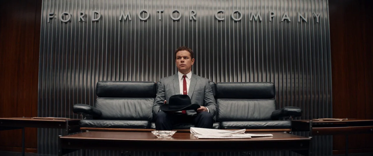Ford V Ferrari (2019) movie still: medium — A modern corporate office features a metal corrugated wall with silver lettering. A man sits centrally…
