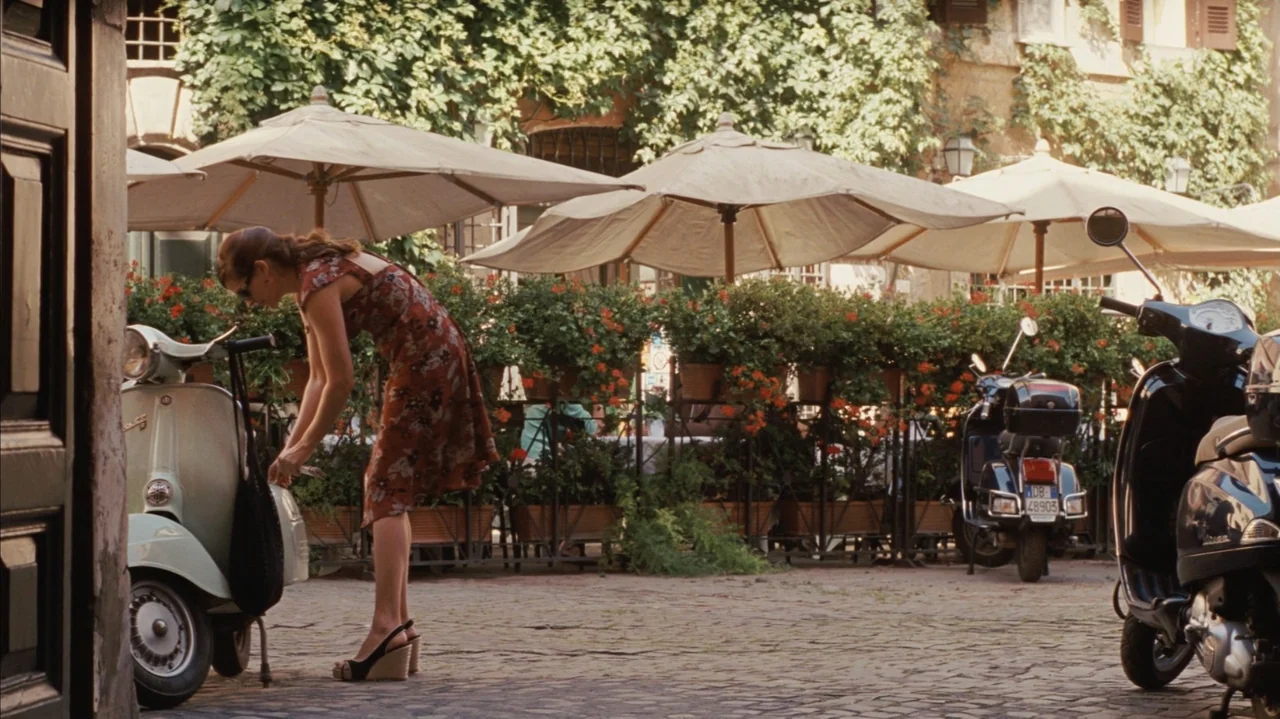 Duplicity (2009) movie still: medium — A sunlit cobblestone plaza features multiple outdoor cafe umbrellas and climbing vines on walls. A woman in a…