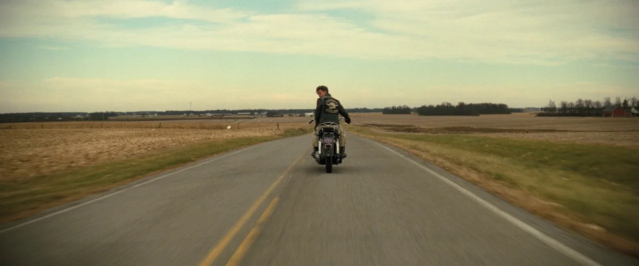 The Bikeriders (2023) movie still: wide — A flat asphalt country road stretches through brown autumn fields under an overcast sky. A man on a…