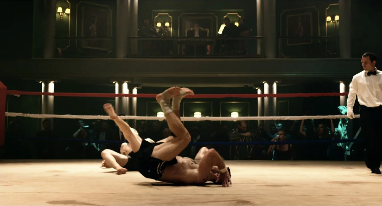 Boyka: Undisputed IV (2016) movie still: fullbody — A well-lit fighting arena features a raised floor with ropes and a spectator balcony. Two men…