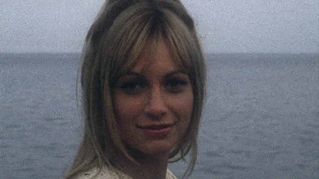Away From Her (2006) movie still: closeup — A woman stands before a large, calm body of water under a gray sky. She looks towards the viewer with a…