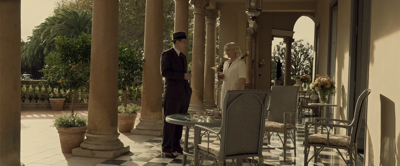 Marlowe (2023) movie still: fullbody — A stone patio with columns and a black-and-white checkered floor overlooks a garden area. A man in a suit and…